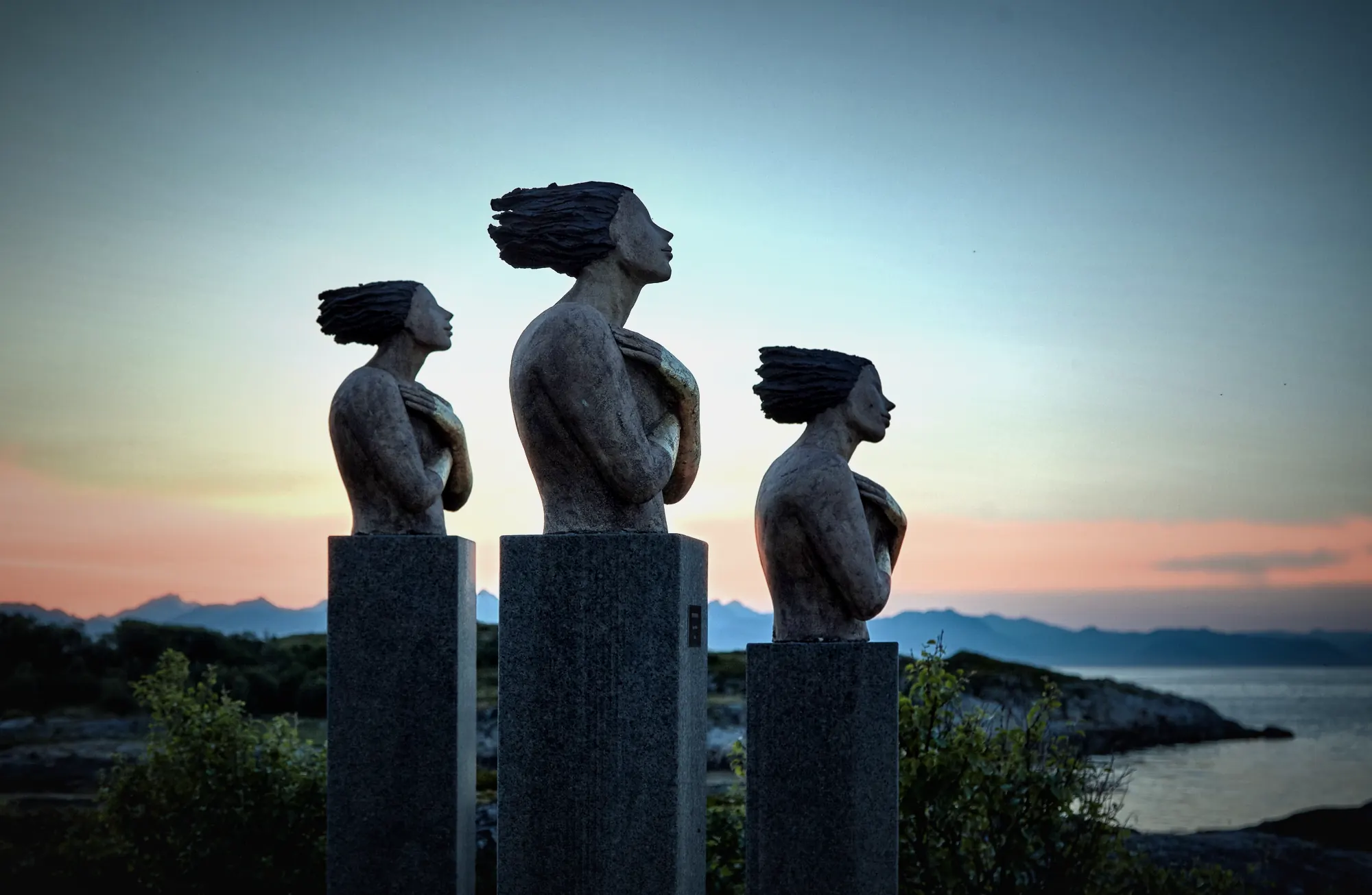 Three sculpted figures on stone plinths, facing the sea at dusk